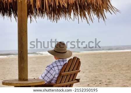 View of a Caucasian man with a hat sitting on an Adirondack chair under a palapa umbrella at the beach.