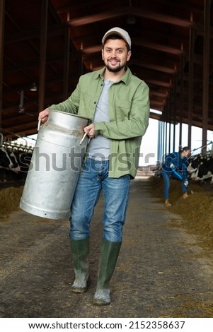 Full length portrait of young bearded farmer working on dairy farm, carrying milk churn near stall with cows in cowshed.. Royalty-Free Stock Photo #2152358637