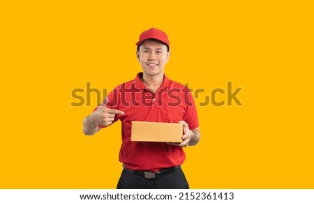 Asian delivery man worker smiling and pointing finger at the box in red uniform isolated on yellow background, hold parcel boxes for sending or conveying parcels by mail. Royalty-Free Stock Photo #2152361413