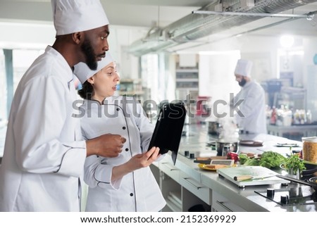Gourmet cuisine experts with modern tablet searching for dinner service dish ideas on internet. Professional chefs with handheld touchscreen device preparing ingredients for evening dinner service.