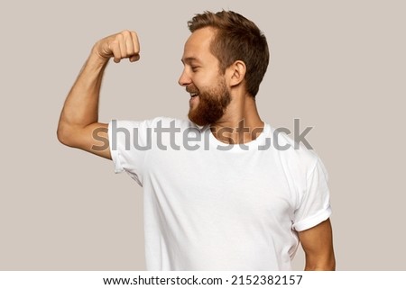 Picture of cheerful guy of 30s dressed in white sport t-shirt with copy space for your advertising content, showing his strong biceps with proud, having red-haired thick beard, isolated over gray wall