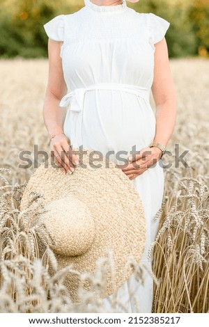 A pregnant young woman in a white long summer dress holding a hat in her hand and her belly