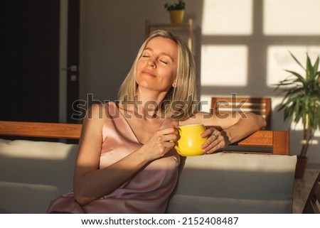 Satisfied middle aged woman is sitting on couch at home with cup of coffee. Smiling adult female of 40 years old with close eyes is sitting on sofa in living room