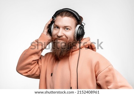 Scandinavian handsome contented man looks up smiles and listens to music in professional headphones isolated on gray background. Happy guy with ginger hairstyle and beard. Modern digital technologies