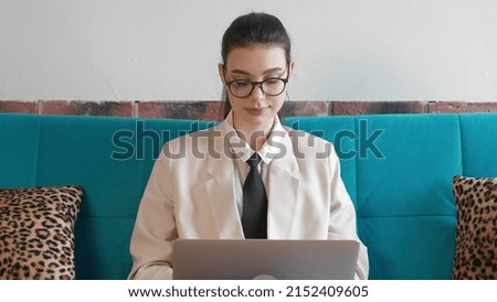 A young freelance woman in a shirt and slippers, working remotely in a home office, uses a laptop while sitting on the couch. A modern woman browses the Internet in search of information, communicates