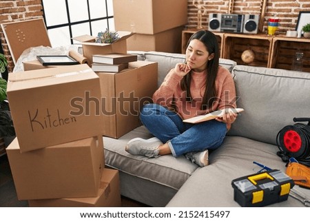 Young hispanic woman with doubt expression sitting on sofa at new home