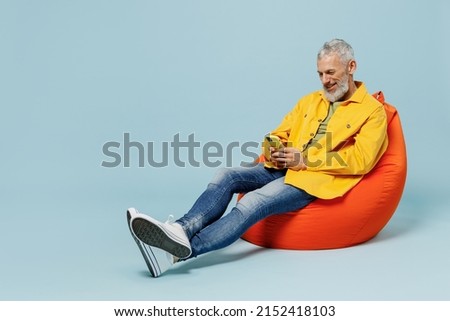 Full body smiling happy elderly gray-haired mustache bearded man 50s in yellow shirt sit in bag chair hold in hand use mobile cell phone isolated on plain pastel light blue background studio portrait.