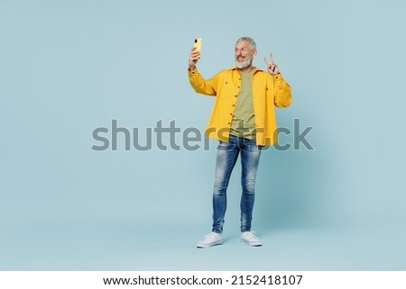 Full body elderly gray-haired mustache bearded man 50s wear yellow shirt doing selfie shot on mobile cell phone post photo on social network show v-sign isolated on plain pastel light blue background