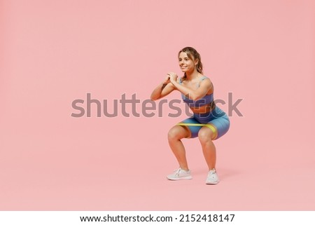 Full body young sporty athletic fitness trainer instructor woman wear blue tracksuit spend time in home gym use fitness rubber bands do squats isolated on plain pink background. Workout sport concept