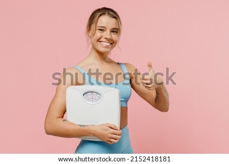 Young happy sporty athletic fitness trainer instructor woman wear blue tracksuit spend time in home gym hold scales show thumb up isolated on pastel plain light pink background. Workout sport concept