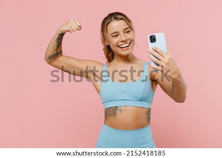 Young strong sporty athletic fitness trainer woman wear blue tracksuit spend time in home gym do selfie shot on mobile cell phone show muscles isolated on plain pink background. Workout sport concept