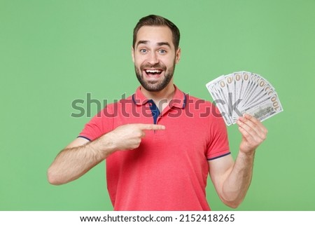 Excited young bearded man guy in casual red pink t-shirt isolated on green background. People lifestyle concept. Mock up copy space. Pointing index finger on fan of cash money in dollar banknotes