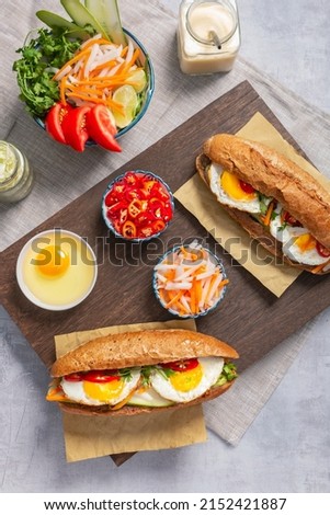 Table top flatlay shot of Banh Mi - Vietnamese sandwich with sausage, pork, lettuce, tomato, eggs and arugula on the light grey texture background. Surrounding by ingredients Royalty-Free Stock Photo #2152421887