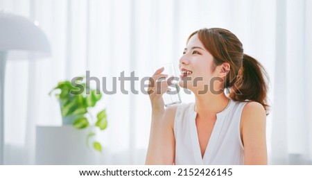 side view of asian woman with brunette ponytail smile drinks a glass water happily in a white room Royalty-Free Stock Photo #2152426145