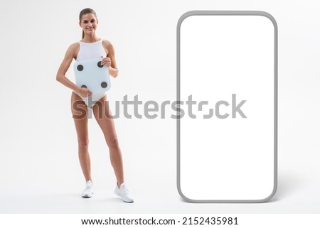 Cute athletic sporty female holding bathroom scales dressed in white swimsuit, smiling standing next to big phone template with copy space screen. Losing weight concept