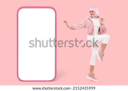 Fancy female in pink wig and fake fur coat dancing over pinkish background next to big banner of cellphone screen for advertising content, presenting new music application Royalty-Free Stock Photo #2152435999
