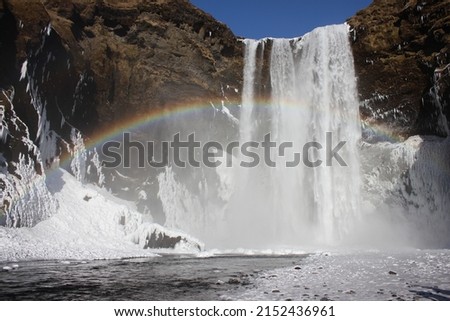 A beautiful full arch rainbow appears during a sunny winter day with lights striking water droplets of the majestic Skogafoss waterfall surrounded by snow and ice in the south of Iceland, Europe