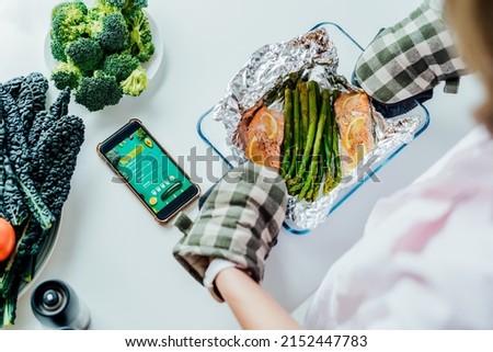 Pescetarian meal diet recipes. Top view woman holding form with baked salmon and asparagus. Phone with Pescetarian menu in mobile lying on the table. Healthy eating and dieting, weight loss concept. Royalty-Free Stock Photo #2152447783