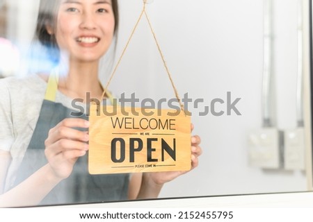 Asian small business owner woman turning shop entrance sign to open again after the quarantine due to coronavirus pandemic. Woman hanging open sign on the glass window. Focus on sign.