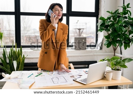 Woman architect talking on smartphone,  working on interior renovation in office. Designer discussing with client visualization and blueprints of the project. Architecture and interior design concept