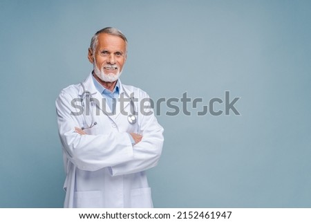 Smiling grey-haired male doctor, physical, therapist wearing white medical gown with stethoscope on shoulders stands with arms crossed isolated on blue, copy space. Healthcare and medicine Royalty-Free Stock Photo #2152461947
