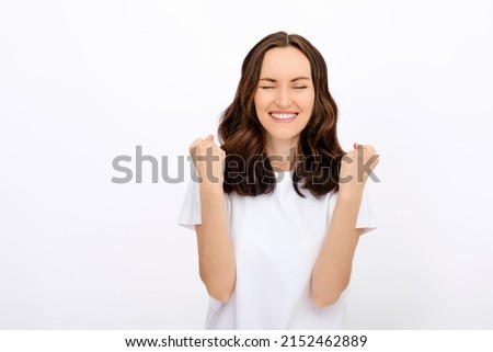 portrait of smiling pleased brunette woman with closed eyes in white T-shirt with bent arms and fists at the top on white background for advertising, shows luck, success concept, copy space, template