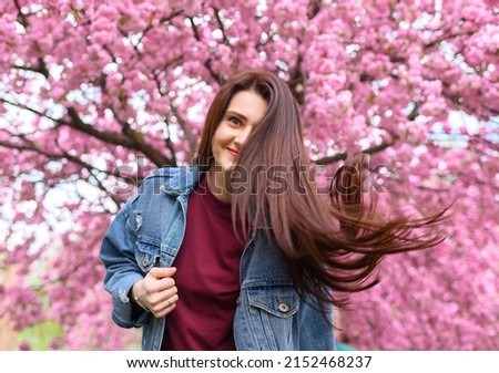 Young beautiful girl in a blue denim jacket is spinning on a background of blooming pink sakura tree and her hair is flying