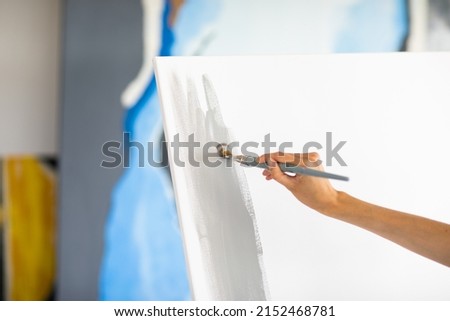 Woman's hand is painting big grey picture stands on the easel at home. Focus is at the brush. Art concept. Royalty-Free Stock Photo #2152468781