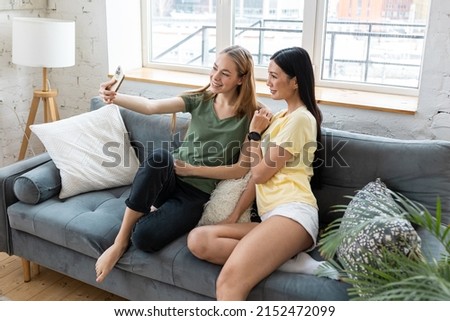 Two millennial diverse girl friends at home watching movies, taking photo, spending fun time together, chat, talk. Females using technology. Asian and Caucasian model.