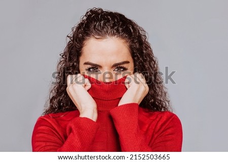 Pretty young woman with curly hair, wearing warm red turtle neck sweater, covers half of her face, on grey background. Studio portrait. Royalty-Free Stock Photo #2152503665
