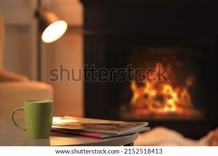 Cup of hot drink and magazines on table near fireplace at home, space for text. Cozy atmosphere