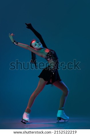 Free dance. Dynamic portrait of young girl, female figure skater in black stage dress skating isolated on blue background in neon light. Concept of movement, sport, beauty. Copy space for ad, text