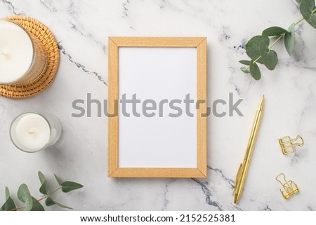 Top view photo of wooden photo frame candles on rattan serving mat gold pen binder clips and eucalyptus on white marble background with empty space