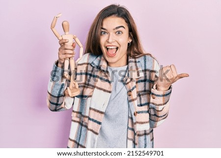 Young caucasian girl holding small wooden manikin pointing thumb up to the side smiling happy with open mouth 