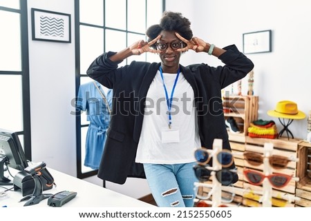 African young woman working as manager at retail boutique doing peace symbol with fingers over face, smiling cheerful showing victory 