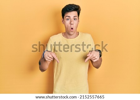 Young hispanic man wearing casual yellow t shirt pointing down with fingers showing advertisement, surprised face and open mouth 