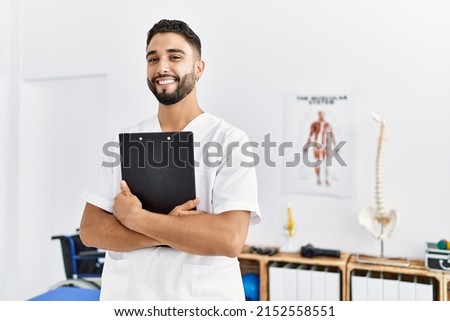 Young arab man wearing physiotherapist uniform holding clipboard at clinic Royalty-Free Stock Photo #2152558551