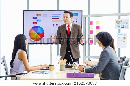 Asian professional successful businessman manager standing showing company growth profit target graph chart presentation on computer monitor to young male and female colleagues in office meeting room.
