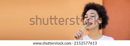 curly and young african american woman eating jelly stick near orange wall, banner