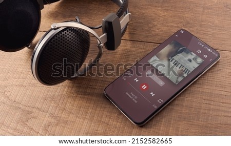 A pair of headphones and a smartphone on a wooden table. On the display you see the player, and a music album playing. Concept of new ways to relax anywhere.