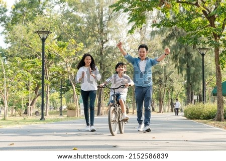 Happy Asian father and mother teach their son to ride a bicycle, Cheerful parent raise hands up in the air to support kid encouragement, family do activity together at park concept Royalty-Free Stock Photo #2152586839