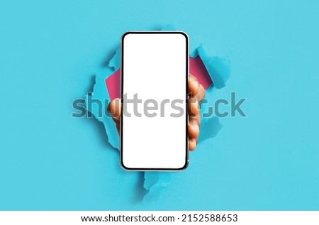 Mobile App, Great Offer. Black male hand holding smartphone with white empty screen showing device close up to camera breaking through blue paper sheet. Gadget display with free copy space, mock up Royalty-Free Stock Photo #2152588653