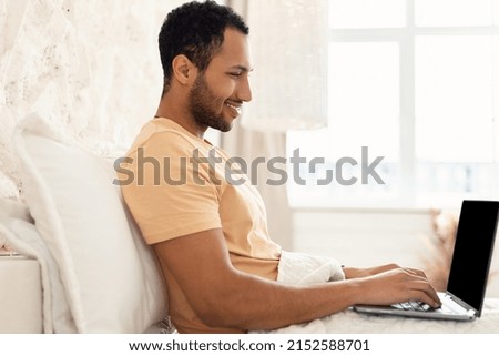 Happy Freelancer Man Using Laptop Computer Typing And Browsing Internet Working Online Sitting In Bed In Modern Bedroom Indoors. Freelance Career And Remote Job Concept