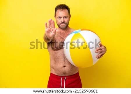 Middle age caucasian man holding beach ball isolated on yellow background making stop gesture