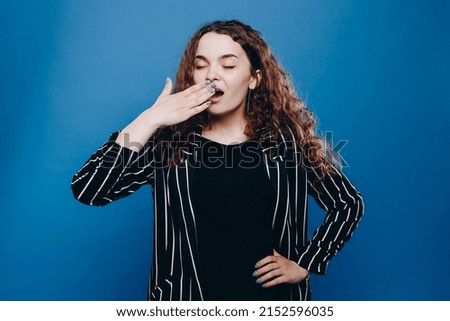 Exhausted sleepless woman has curly hair yawns covers mouth in wants to sleep awakes very early isolated over blue background. People sleeping disorders concept