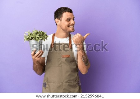 Brazilian Gardener man holding a plant over isolated purple background pointing to the side to present a product