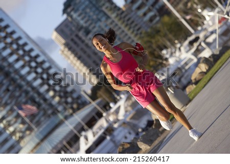 USA, California, San Diego, woman wearing pink sports vest and shorts, jogging, listening to MP3 player strapped to arm, smiling, marina in background (tilt)