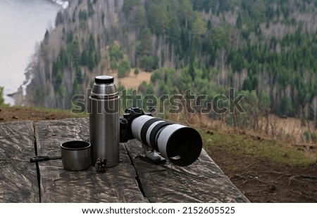 on a wooden table in nature there is a thermos a camera with a telephoto  lens