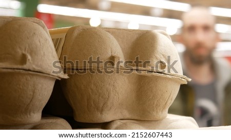 Close-up of a cardboard packaging of eggs on a store shelf and a blurry background with a male customer behind it