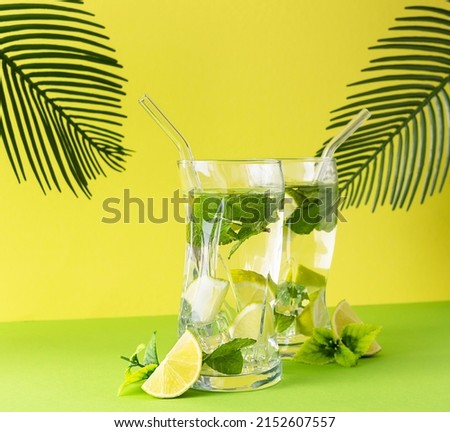 Two transparent glasses of mojito on a green table. Palm leaves in the background. Dalki lajama and mint leaves on the table. Yellow background